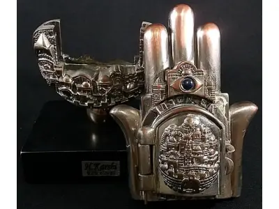 LOTE 285 - KARSHI RARO GRUPO ESCULTÓRICO EM METAL ESPESSURADO A PRATA DE LEI 925 MILÉSIMOS, CONTRASTADA NA BASE E SELADA, REPRESENTA HAMSÁ, COM CIDADE DE JERUSALÉM EM RELEVO NA PARTE CENTRAL, ONDE SE ABRE A PORTA PARA ACESSO A PÁGINA DO ALCORÃO., 21 CM DE ALT.(NO ESTADO)