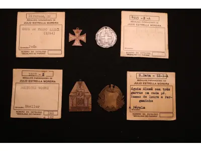 LOTE 036 - LOTE 36 – MEDALHAS ALEMANHA 	MEDALHA	OBSERVAÇÕES 1.		Cruz de Ferro Alemã. 1914 Gravador: José Peón	Reverso liso - envelope da Coleção Júlio Estrella Moreira 2.		Beneficente da Cruz Vermelha Austro-Alemã. 1915	Prateada - envelope da Coleção Júlio Estrella Moreira 3.		Deustche Woche (Semana Alemã). 1937. Curitiba Gravador: Eduardo Muller	Reverso liso - envelope da Coleção Júlio Estrella Moreira 4.		Águia Alemã com pergaminho Gravador: Henrique Kaminski – A Pérola	Reverso liso - envelope da Coleção Júlio Estrella Moreira