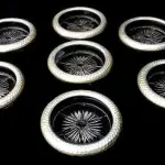 LOTE 142 - CONTEMPORÂNEO JOGO COM 7 PORTA COPOS EM VIDRO TRANSLÚCIDO COM BASE ESTRELADA E BORDA ADORNADA EM ALUMÍNIO MARTELADO. MED 9CM DIÂM X 1,5CM ALT (NO ESTADO)