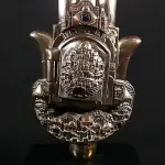 LOTE 285 - KARSHI RARO GRUPO ESCULTÓRICO EM METAL ESPESSURADO A PRATA DE LEI 925 MILÉSIMOS, CONTRASTADA NA BASE E SELADA, REPRESENTA HAMSÁ, COM CIDADE DE JERUSALÉM EM RELEVO NA PARTE CENTRAL, ONDE SE ABRE A PORTA PARA ACESSO A PÁGINA DO ALCORÃO., 21 CM DE ALT.(NO ESTADO)