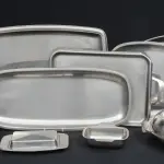 LOTE 297 - SERVIÇO DE MESA COMPLETO COM 14 PEÇAS DE AÇO INOX: 6 BANDEJAS  4 FORMAS REDONDAS; 1 FORMINHA; 2 PRATOS E UMA MANTEIGUEIRA.(NO ESTADO)