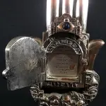 LOTE 285 - KARSHI RARO GRUPO ESCULTÓRICO EM METAL ESPESSURADO A PRATA DE LEI 925 MILÉSIMOS, CONTRASTADA NA BASE E SELADA, REPRESENTA HAMSÁ, COM CIDADE DE JERUSALÉM EM RELEVO NA PARTE CENTRAL, ONDE SE ABRE A PORTA PARA ACESSO A PÁGINA DO ALCORÃO., 21 CM DE ALT.(NO ESTADO)